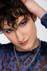portrait of nonbinary person with makeup and silver necklaces touching wavy hair isolated on grey.