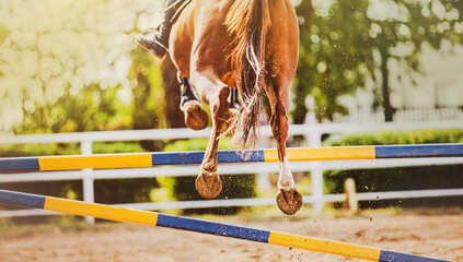 The hooves of a beautiful sorrel horse jumping over a high yellow-blue barrier on a sunny summer...