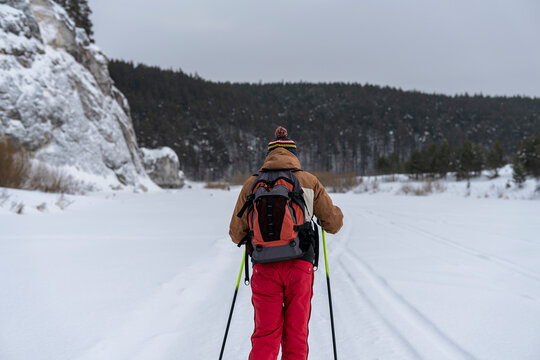 Rear View Of Young Man In Red Brown Clothes With Backpack Skiing Near Rocks And Cliffs Active Healthy Lifestyle Winter Sports Hiking