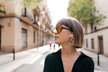 Close up profile portrait of stylish young woman with short hair wearing black shirt listens music on city background in sunlight. Mood, lifestyle, concept.