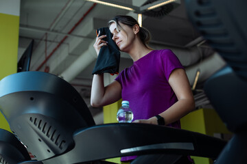 Overweight tired woman run on treadmill at fitness gym wiping sweat from face by towel, side view....