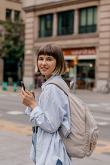 Lovely stylish appealing girl with short hair wearing blue shirt with backpack walking in the city with smartphone 