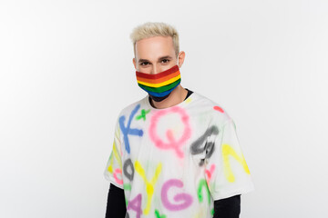 blonde gay man in trendy t-shirt and lgbtq colors medical mask looking at camera isolated on grey.
