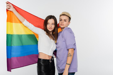 tattooed lesbian woman looking at camera near girlfriend with lgbtq flag isolated on grey.