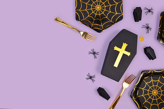 Halloween party flat lay with spider web plates, coffins, confetti and spiders on violet background with copy space