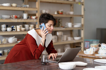 Young creative woman artisan artist ordering materials for ceramic work by phone while working in studio or workshop, female pottery shop owner talking with client via cellphone and typing on laptop