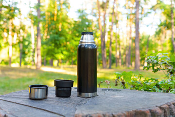 Black thermos bottle and cups on a tree stump - obrazy, fototapety, plakaty