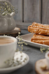 A white cup of coffee with milk stands on a gray background, behind a plate with eclairs