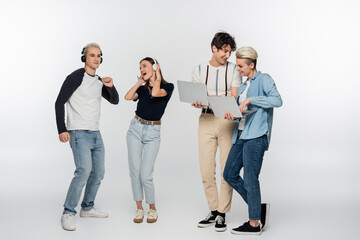 Fototapeta premium smiling couple using laptops near cheerful friends dancing in wireless headphones on grey background.