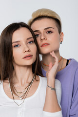 brunette lesbian woman touching chin of blonde girlfriend while looking at camera isolated on grey.