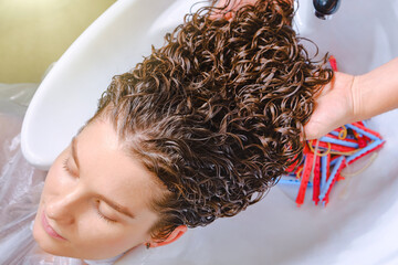 Professional hairstylist washes client’s hair with conditioner shampoo for washing after curling hair. Close up of curlers in hair. Creating hairstyles by a hairdresser in the salon. Shallow depth - obrazy, fototapety, plakaty