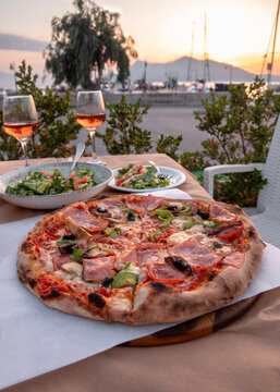Delicious pizza with cheese, bacon, salami, mushrooms and green pepper and two glasses of wine against the background of the port in Greece