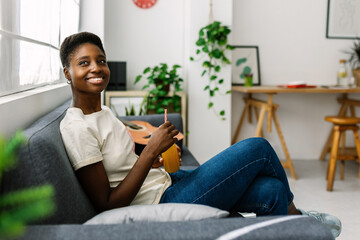 Young african woman chilling on sofa drinking orange juice - Millennial dreamy female relaxing at...