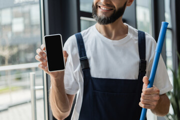 partial view of happy bearded cleaner in overalls holding mobile phone with blank screen.