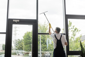 back view of man in overalls washing large office windows with window squeegee.