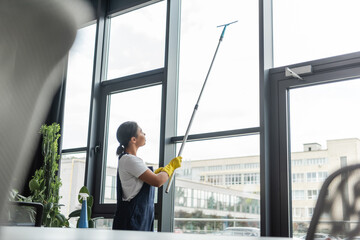 bi-racial woman in workwear washing large office windows on blurred foreground.