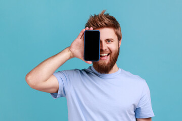 Portrait of smiling optimistic bearded man covering half face with cellphone and looking at camera with toothy smile, positive glad face. Indoor studio shot isolated on blue background. - Powered by Adobe