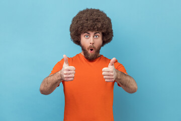 Shocked man with Afro hairstyle wearing orange T-shirt showing thumbs up, looking at camera with...