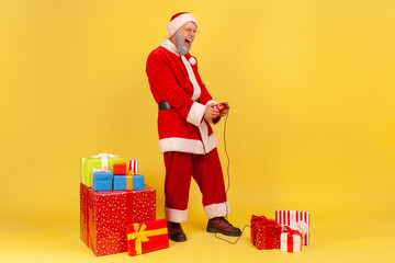 Full length portrait of elderly man with gray beard in santa claus costume standing with joystick, playing video game, surrounded with Christmas gifts. Indoor studio shot isolated on yellow background © khosrork