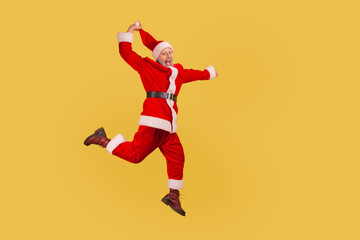 Fototapeta na wymiar Full length of elderly man with gray beard wearing santa claus costume jumping up high and pulling his red hat, celebrating Christmas with excitement. Indoor studio shot isolated on yellow background.