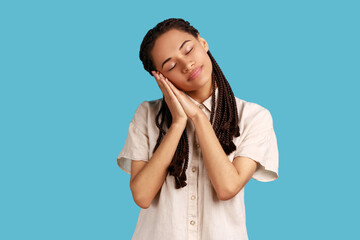 Portrait of beautiful woman with black dreadlocks sleeping laying down on her palms, having comfortable nap and resting, dozing off, wearing white shirt. Indoor studio shot isolated on blue background