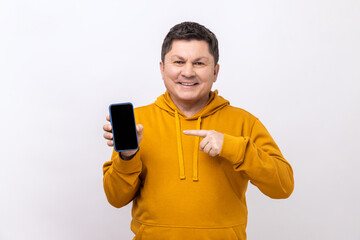 Middle aged man pointing finger at smartphone with empty screen, looking at camera with smile, freespace for advertisement, wearing urban style hoodie. Indoor studio shot isolated on white background.
