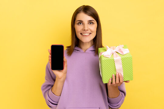 Present, bonus for mobile user. Portrait of attractive young adult woman holding gift box and cell phone with mock up, blank display, wearing hoodie. Indoor studio shot isolated on yellow background.