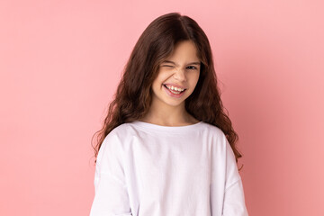 Portrait of playful cheerful little girl wearing white T-shirt blinking eye as having cunning idea, winking and smiling to camera. Indoor studio shot isolated on pink background.
