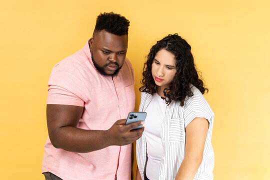 Portrait Of Serious Concentrated Young Couple In Casual Clothing Standing Together, Using Mobile Phone, Looking At Display, Checking Social Networks. Indoor Studio Shot Isolated On Yellow Background.