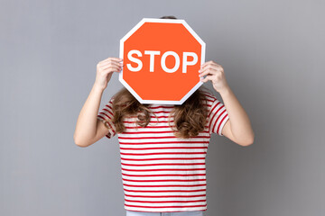 Little girl in striped T-shirt covering face with Stop symbol, anonymous woman holding red traffic...