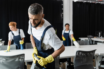 man with floor scrubber machine near interracial women cleaning desks on blurred background.