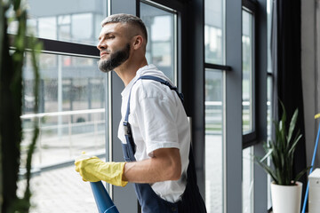 bearded professional cleaner in overalls looking through window in office.