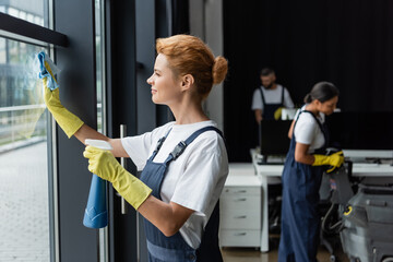woman in rubber gloves cleaning window with rag and detergent near blurred interracial colleagues.