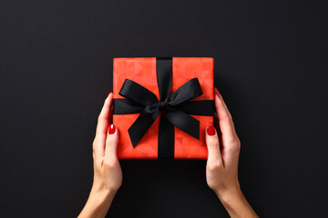 Luxury red gift box with black ribbon bow in female hands over black background. Black Friday sale gift, premium present for Birthday, Christmas, Valentines day