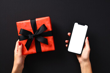 Black Friday sale concept. Female hands holding mobile phone mockup with blank screen and red gift box over black background. Black Friday online shopping. - Powered by Adobe