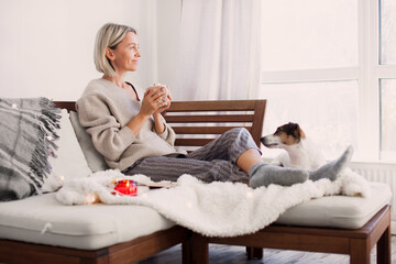 Happy dreamy middle aged woman sitting on sofa in living room with cup of tea or coffee