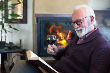Senior man drinking wine and reading book by fireplace in winter
