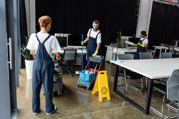 woman in overalls with floor scrubber machine near man with cart of cleaning supplies.