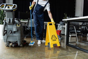 cropped view of man with mop holding caution sign board near floor scrubber machine.