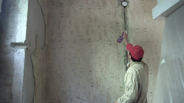 Caucasian builder in uniform is moistening old wallpapers in apartment room to rip it off. Slowmo of man doing repairs in his house. He wants to change interior and coverage in flat. Home improvment