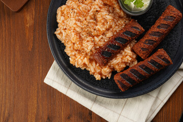 Traditional south european skinless sausages cevapcici made of ground meat and spices on black plate on dark wooden board, vegan substitute made of soy protein, with rice and sour cream, top view