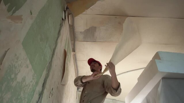 Process of removing old wallpaper from ceiling by man in uniform standing on ladder in room filmed in slow motion from below. Young adult caucasian builder is doing repairs in his house. Home renewal.