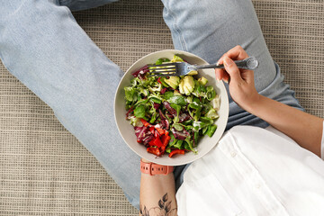 Female with tattoos wearing blue jeans and white shirt sitting and holding a grey bowl with salad...