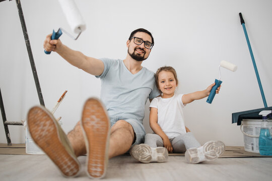 Cheerful caucasian man father and his cute little girl daughter sitting on floor in white light room background with paint rollers in their hands.