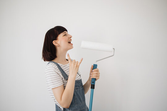 Young beautiful woman with red hair singing song at new home, girl using into a mop and a paint roller instead of real microphone showing rock sign