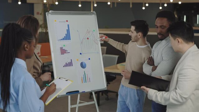 Smart male manager presenting analytical report on flip chart during conference with coworkers at office. Multiracial colleagues standing near and taking notes during meeting.