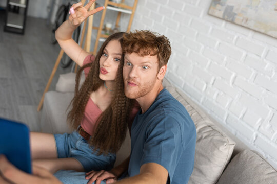young redhead man taking selfie with girlfriend pouting lips and showing peace sign in living room.