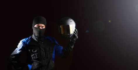 male motorcyclist in a motorcycle outfit and a balaclava puts on a helmet on a dark background. The...