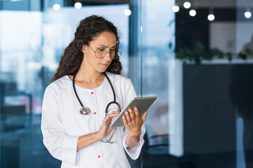 Serious thinking hispanic female doctor using tablet computer, female doctor in glasses and white medical coat with stathoscope working inside office clinic.