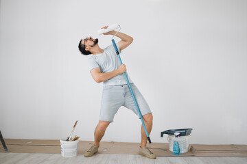 Happy caucasian male singing song using roller brush as a microphone. Young man having fun during home repair and renovation